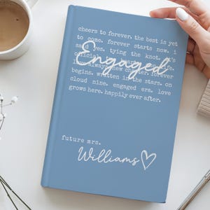 Puede incluir: Un diario azul claro con la palabra "Engaged" en el centro y el nombre "Williams" debajo. El diario tiene texto que dice "cheers to forever" y "future mrs."