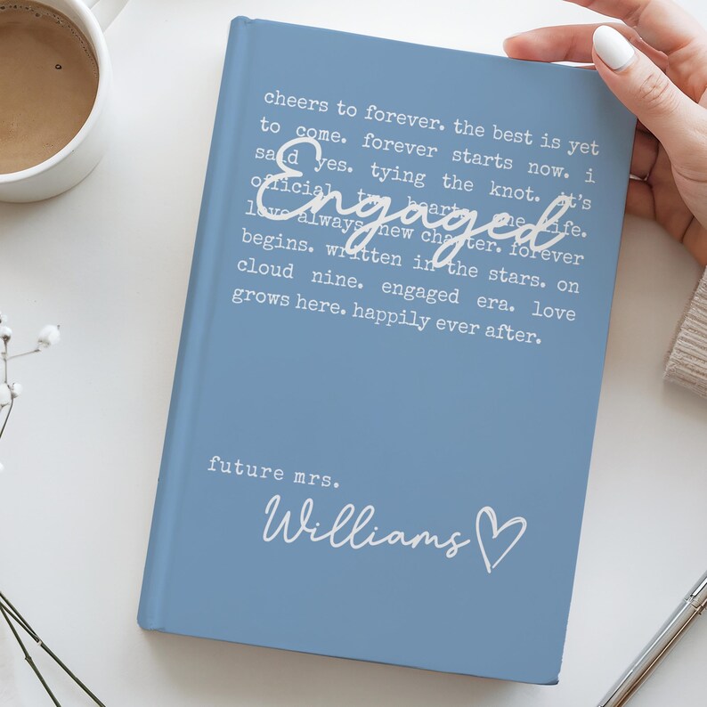 Puede incluir: Un diario azul claro con la palabra "Engaged" en el centro y el nombre "Williams" debajo. El diario tiene texto que dice "cheers to forever" y "future mrs."