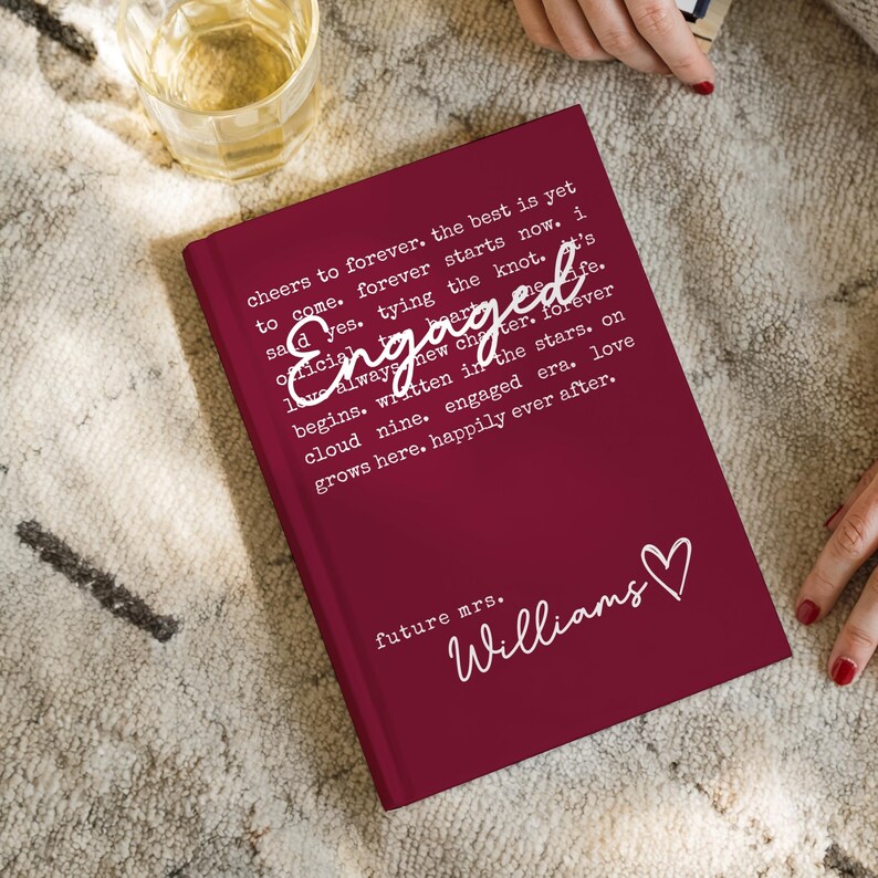 Puede incluir: Un diario de color burdeos con la palabra "Engaged" en el centro. El diario tiene texto blanco que dice "cheers to forever" y "future mrs. Williams" con un s&iacute;mbolo de coraz&oacute;n. Un vaso de l&iacute;quido est&aacute; en la esquina superior izquierda.