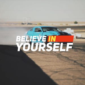 May include: A blue sports car drifts on a racetrack, with the text "BELIEVE IN YOURSELF" overlaid. A person in an orange vest is visible on the left, possibly taking photos. The word "KINETIC" is in the upper left corner.