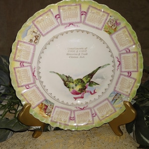 May include: A decorative plate with a scalloped edge, featuring a central image of two birds and text that reads "Compliments of COOK & COOK Groceries & Fruit Clinton, Ind." The plate has a green and yellow border with calendar-like images.