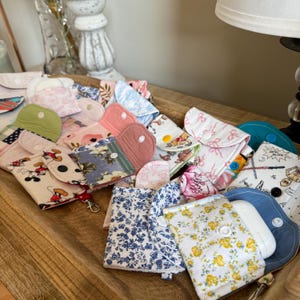 May include: A collection of handmade fabric pouches in various colors and patterns, including floral, Mickey Mouse, and solid colors. Each pouch has a button closure. Some pouches are open, revealing their contents.