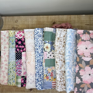 May include: A collection of folded fabric pieces with various floral and patterned designs. The fabrics include pink bows, blue flowers, patchwork, and other colorful prints. The fabrics are arranged on a wooden surface.