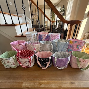 May include: A collection of handmade fabric tote bags in various sizes and patterns. The bags feature diverse designs, including polka dots, floral prints, and celestial motifs. The color palette includes pinks, purples, and greens, with contrasting interior linings.