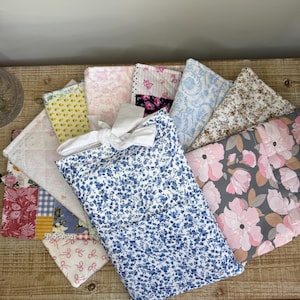 May include: A collection of quilted fabric pouches in various floral and patterned designs. The pouches feature a range of colors, including blue, pink, yellow, and white. One pouch is tied with a white bow. The fabrics appear to be cotton.