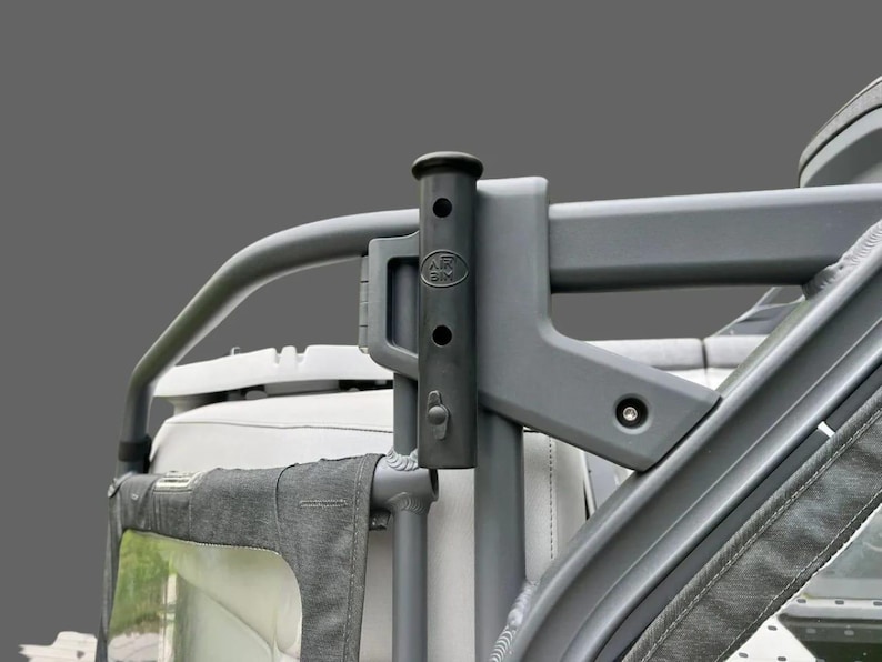 May include: A gray metal roll bar with a black vertical support. The support has a cylindrical top and several holes. The letters "AK BIX" are visible on the support. The roll bar is attached to a vehicle.