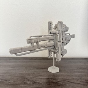 May include: A grey model of a futuristic weapon or spacecraft component. The design features a complex arrangement of rectangular and geometric shapes, supported by a thin stand. The object is displayed against a neutral background.