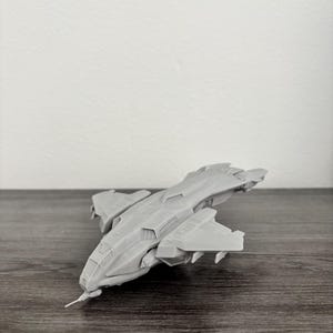 May include: A gray model spaceship with a detailed design, featuring wings and a pointed front. The spacecraft is positioned on a wooden surface, with a white wall in the background. The model appears to be a miniature replica of a futuristic aircraft.