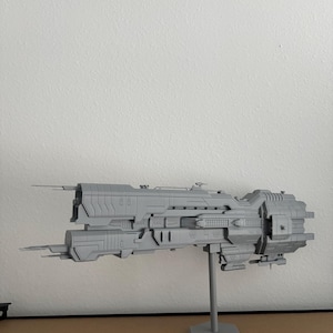 May include: A grey model spaceship with a detailed, futuristic design. The model is mounted on a grey stand, displaying its long, rectangular body with multiple protrusions and a detailed surface. The spaceship is a model of a science fiction vehicle, suitable for display.
