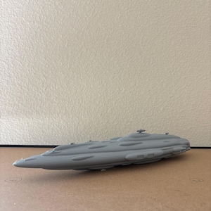 May include: A gray, 3D-printed model of a spaceship. The model has a long, streamlined body with several raised sections and small details. The spaceship is resting on a brown surface.
