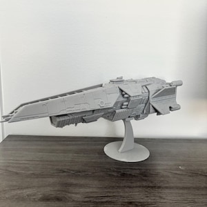 May include: A gray model spaceship with intricate details, mounted on a circular stand. The spaceship has a long, pointed front and various structural elements. The model is made of a solid, light gray material and is displayed against a neutral background.