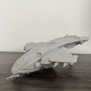 May include: A gray model spaceship with a detailed design, featuring wings, a cockpit, and various exterior components. The spacecraft is positioned on a dark wooden surface, with a plain white background.