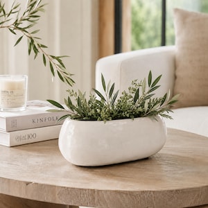 May include: An elongated, smooth, white ceramic planter bowl filled with green foliage. The planter sits next to two stacked books, one labelled Kinfolk and the other labelled at 100, and a glass candle jar with a label reading Santal 26.