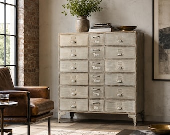 May include: A sixteen-drawer metal cabinet with a distressed, light grey finish. Each drawer is labelled with a number from one to six. A vase of greenery sits on top. The cabinet has metal handles and is in a room with a leather armchair and a brick wall.
