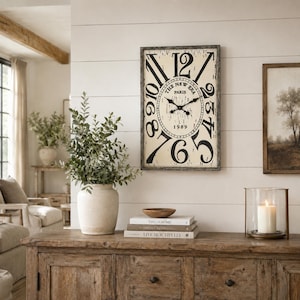 May include: A rectangular wall clock with a distressed cream face, black Roman numerals, and the text THE NEW ERA PARIS 1989. The clock features a dark wooden frame, black metal hands, and a weathered finish, mounted on a white horizontal plank wall.