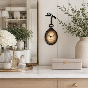 May include: An oval wall clock with a dark brown beaded frame and a cream-colored face featuring black Roman numerals and hands. The clock is suspended by a black ribbon tied in a bow at the top, creating a pocket watch aesthetic on the wall.