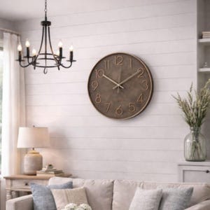 May include: A round, bronze-colored wall clock with raised numerals and hands. The clock is mounted on a white, horizontal plank wall. The word "QUARTZ" is printed on the clock face. A black chandelier hangs from the ceiling. A beige sofa and pillows are in the foreground.