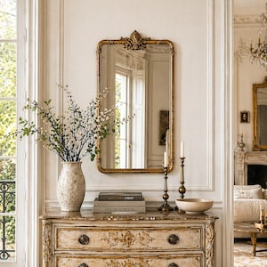 May include: A gold-framed rectangular mirror with ornate detailing hangs above a vintage chest of drawers. A vase of flowers, books, candles, and a bowl sit on the chest. The room has a classic, elegant aesthetic.