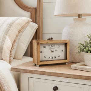 May include: A rectangular wooden table clock with a white face, black numerals, and a small metal handle on top. The clock face features the text Grand Hotel and 1850. It stands on four small silver-toned feet and has black hour and minute hands.