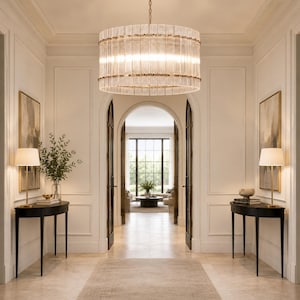 May include: A large cylindrical chandelier featuring vertical glass rods arranged in a drum shape with gold-toned metal bands at the top, middle, and bottom, suspended from a chain in a bright, symmetrical hallway with white walls.