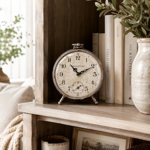 May include: A round, vintage-style analog clock with a distressed metal frame and a cream-colored face. The clock features black Arabic numerals, ornate black hands, and the brand name Bennatyne printed in the center. It has a small sub-dial.