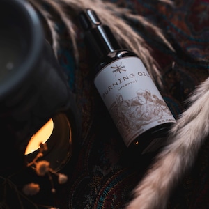 May include: A black glass bottle of "Burning Oil Oriental" with a white label featuring an illustration. The bottle is positioned near a lit candle and decorative dried plants, suggesting aromatherapy or home fragrance.