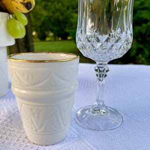 Könnte beinhalten: Eine weiße Keramiktasse mit goldenem Rand und geprägtem Design, neben einem klaren Kristall-Weinglas. Das Weinglas hat ein Rautenmuster auf der Schale und einen langen Stiel. Der Hintergrund ist eine verschwommene Szene im Freien.