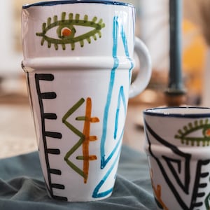 Puede incluir: Taza de cerámica blanca con borde y asa azules, adornada con diseños abstractos pintados a mano en negro, verde, naranja y azul. La taza presenta un motivo de ojo y patrones geométricos.