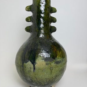 Puede incluir: Un jarrón de cerámica verde, alto, con base bulbosa y cuello cilíndrico. Presenta cuatro protuberancias decorativas y un acabado brillante y texturizado, con tonos verde oscuro y oliva. El jarrón mide aproximadamente 30 cm de altura.