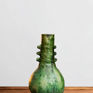 Peut inclure: Un vase en céramique vert de forme bulbeuse avec une surface texturée et marbrée, doté d'un col étroit. Le col présente trois petites languettes horizontales saillantes de chaque côté. La glaçure montre des nuances de vert foncé et clair.
