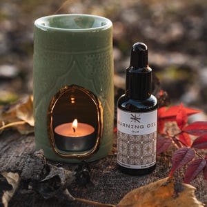 Op de afbeelding: Een groene keramische oliebrander met een goudkleurige opening, met een brandende theelichtkaars. Een zwarte fles "Burning Oil Oud" staat ernaast. De scène speelt zich buiten af met herfstbladeren.