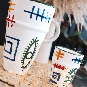 Può includere: Set di brocca e tazza in ceramica bianca con disegni dipinti a mano. La brocca ha un manico ed è decorata con forme geometriche blu, arancioni e rosse. La tazza presenta disegni simili. I bordi sono verdi.
