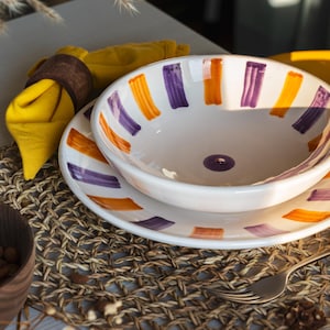 May include: A white ceramic bowl and plate set with orange and purple brushstroke designs. The bowl and plate are on a woven placemat, with a yellow napkin and a wooden bowl of almonds in the background. A fork is also visible.