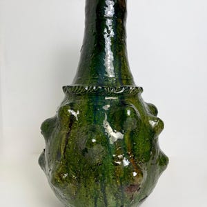 Op de afbeelding: Een handgemaakte, groene keramische vaas met een getextureerd, bolvormig lichaam en een lange, slanke hals. De vaas heeft een glanzende afwerking met kleurvariaties, waaronder donkergroene en bruine tinten. De basis heeft afgeronde uitsteeksels.