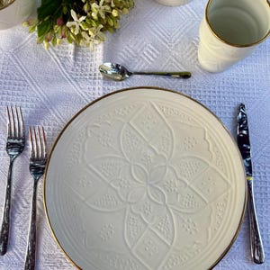 Peut inclure: Une table dressée avec une assiette blanche ornée d'un motif floral et d'un liseré doré. Les couverts en argent comprennent un couteau, deux fourchettes et une cuillère. Une tasse blanche avec une bordure dorée et un petit vase avec des fleurs sont également présents.