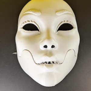 Puede incluir: Una máscara blanca con un rostro humano estilizado. La máscara tiene agujeros negros para los ojos, una nariz pequeña y una boca con dientes visibles. La máscara tiene una superficie lisa y probablemente está hecha de un material duro.