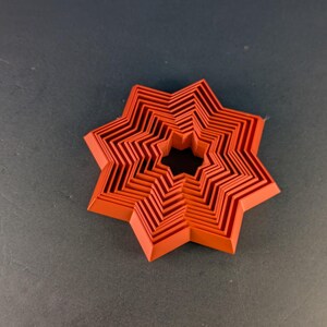Könnte beinhalten: Ein geschichtetes, sternförmiges Objekt in Orange- und Rottönen. Das Objekt hat ein zentrales Loch und besteht aus mehreren Schichten, wodurch ein dreidimensionaler Effekt entsteht. Das Design ist geometrisch und symmetrisch, mit einer modernen Ästhetik.