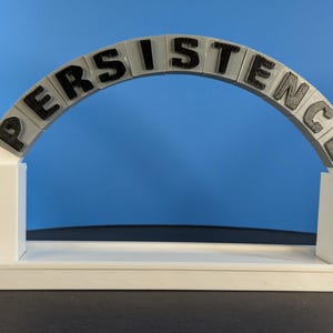 May include: A white 3D-printed arch displaying the word "PERSISTENCE" in black lettering. The arch is supported by two white rectangular pillars and rests on a white base. The background is a solid blue.