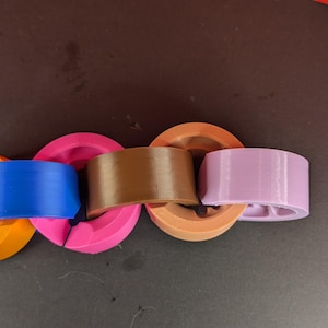 Może przedstawiać: Łańcuch zazębiających się, drukowanych w 3D pierścieni w różnych kolorach: pomarańczowym, niebieskim, różowym, brązowym i lawendowym. Pierścienie są połączone liniowo, prezentując prosty, geometryczny wzór. Obiekty są z tworzywa sztucznego.