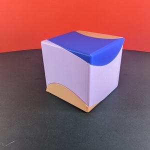 Puede incluir: Un cubo geométrico con un cuerpo lavanda, una parte superior azul y detalles curvos de color dorado. El cubo está sobre un fondo rojo.