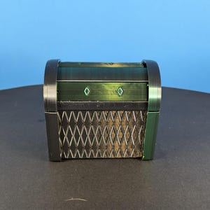 Puede incluir: Un pequeño cofre decorativo con acabado verde oscuro y plateado. El cofre presenta un diseño de celosía en la parte delantera y dos detalles en forma de diamante. La parte superior es arqueada y los lados son de color verde oscuro.