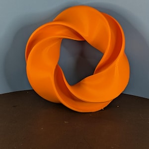 May include: An orange, abstract sculpture with a twisted, circular design. The object is made of a solid material and sits on a dark surface against a blue wall. The sculpture's form creates a continuous loop.