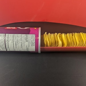 以下が含まれることがあります： ポテトチップスの部分的に開いた円筒形の容器。赤いプラスチック製のトレイにチップスが入っています。容器は紫で、白い文字と栄養成分表示があります。チップスは黄色で、きちんと並んでいます。