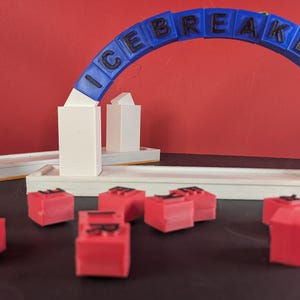 Może przedstawiać: Gra wydrukowana w 3D z niebieskim łukiem z napisem "ICE BREAKERS". Gra zawiera czerwone kostki z literami i białe konstrukcje. Tło jest jednolicie czerwone.