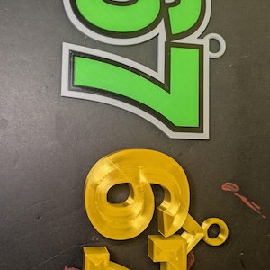 May include: Two 3D-printed number sets, '9' and '7'. The top set is green with a white and black outline. The bottom set is gold. Both sets have a small loop attached. The numbers are on a dark surface.