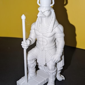 May include: A white statue of the Egyptian god Horus, depicted with a falcon head and a staff. The figure is kneeling on a rectangular base, with detailed carvings and a regal headdress. The statue is set against a yellow background.