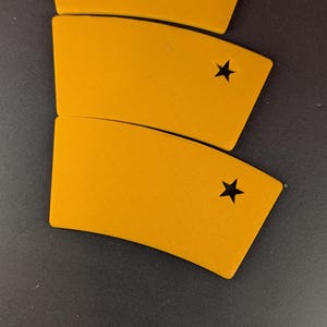 May include: Three mustard-yellow, curved, rectangular objects with a black star cut out of each. The objects are arranged in a slightly overlapping pattern on a dark surface. The objects have a smooth texture.