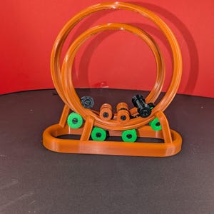 May include: A 3D-printed toy roller coaster in shades of orange, black, and green. The structure features two circular loops and several small cars. The base has green wheels. The background is red.