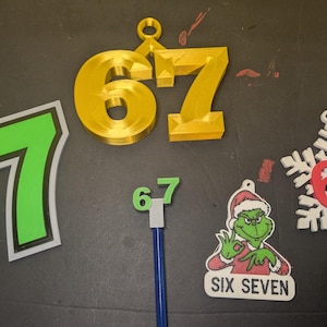 May include: Various decorative ornaments and figures displaying the number "67". Includes a gold 3D-printed number, a green and white number, a Grinch-themed ornament with "SIX SEVEN" text, and a snowflake ornament.
