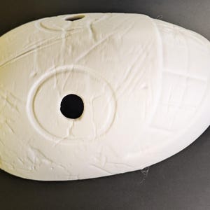 May include: A white, oval-shaped mask with a large central hole and a smaller hole at the top. The mask has a textured surface with circular and linear patterns. The object is likely a decorative piece or a prop.